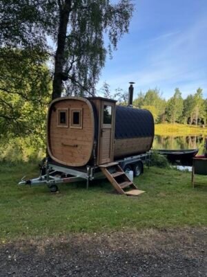 Sidevisning av TimberIN Nordic Square mobilbadstue på tilhenger med skorstein ved en innsjø
